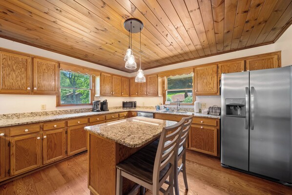 A kitchen with wooden cabinets, granite countertops, a stainless steel refrigerator, and a hanging light fixture. - Riverbank Retreat - Hot Tub, Game Room, & Fantastic River Access (Jefferson)