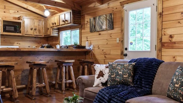 Interior acogedor de una cabaña con paredes de madera, una zona de cocina, una sala de estar con sofá y taburetes de bar, y una puerta con ventanal.