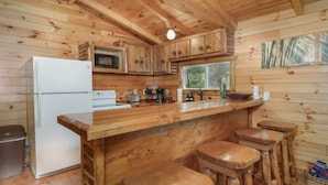 Interior de una cabaña de madera con cocina, que incluye un refrigerador, microondas y taburetes de bar.
