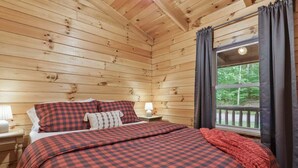 Un dormitorio de cabaña acogedora con ropa de cama de cuadros, una mesita de noche con lámpara y una ventana con cortinas.
