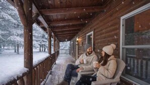 Una cabaña de madera con un porche, dos personas sentadas en un banco y nieve en el suelo.