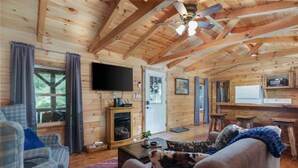 Interior acogedor de una cabaña con paredes de madera, chimenea, televisor y zona de cocina.