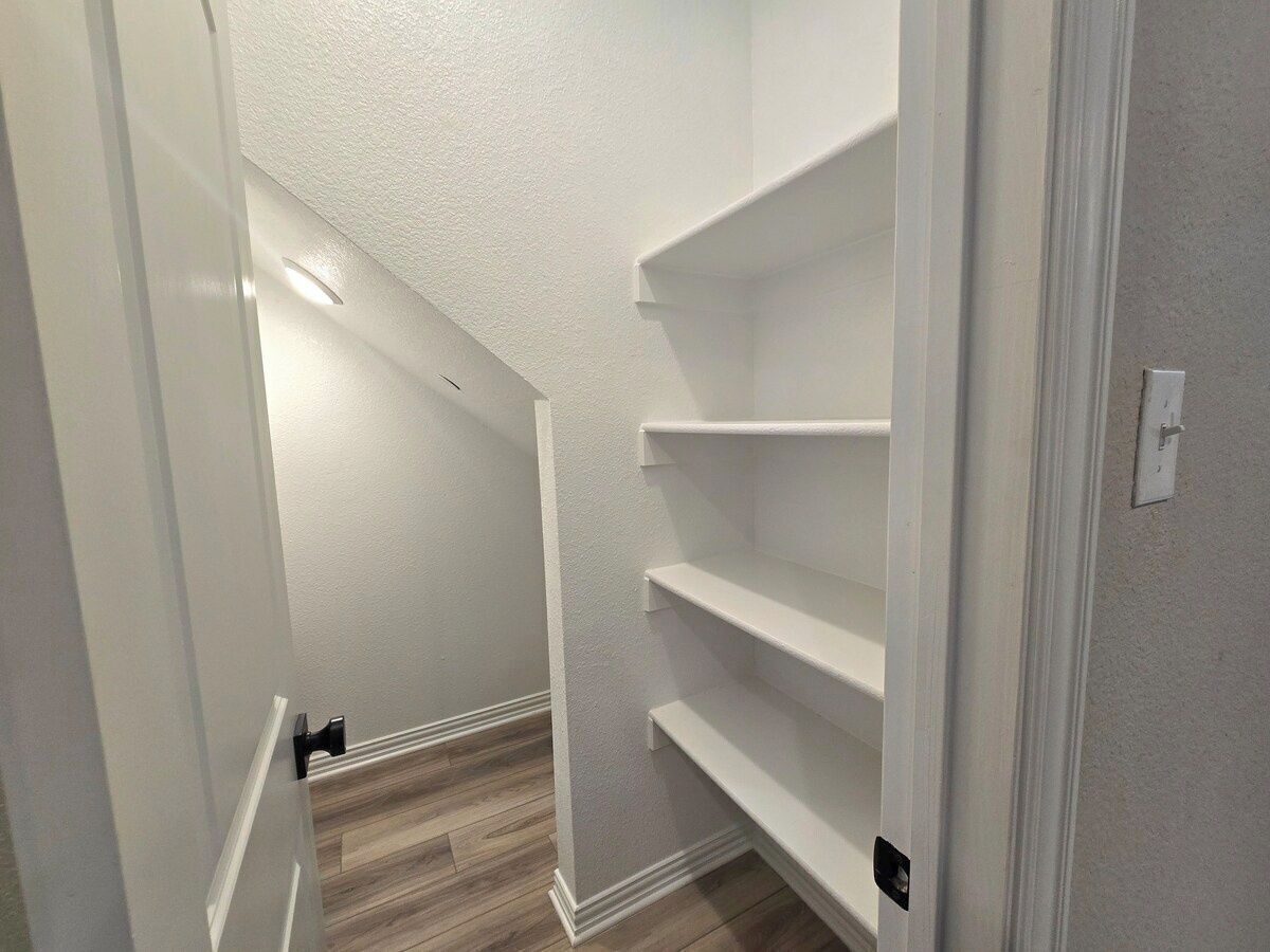 Een smalle gang met witte wanden en houten vloer, die uitkomt bij een trap. Er zijn planken aan de wand bevestigd, en een lamp is zichtbaar aan het plafond.