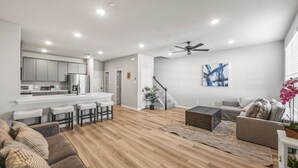 Un salon moderne avec un plancher en bois, un canapé, une table basse et un coin cuisine doté de meubles blancs et d’un bar.