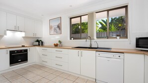 A modern kitchen with white cabinets, a black oven, a dishwasher, and a wooden countertop. There are two windows with a view of greenery outside.