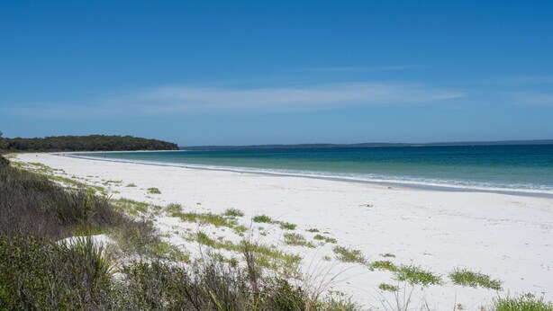 A wide, white sandy beach with clear blue waters and a distant tree-covered landmass.
