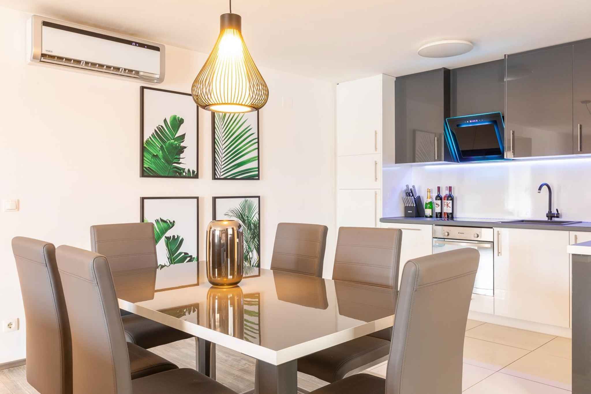 A modern dining area with a table and chairs, a pendant light, framed plant prints on the wall, and a kitchen with a built-in microwave and wine bottles.