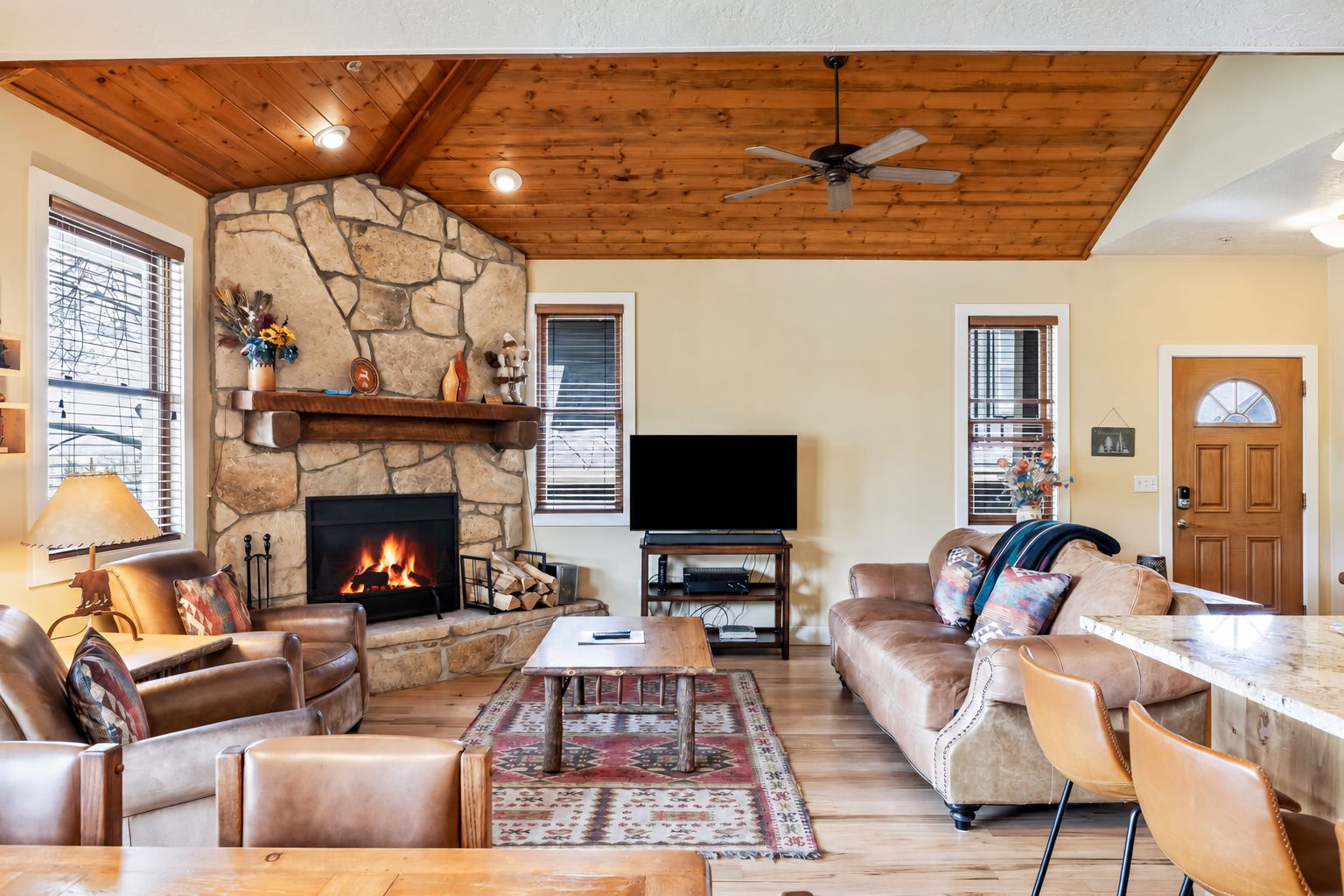 A cozy living room with a stone fireplace, leather furniture, a flat-screen TV, and a ceiling fan.