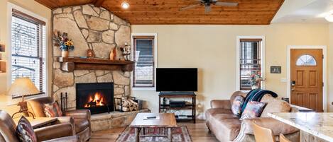 A cozy living room with a stone fireplace, leather furniture, a flat-screen TV, and a ceiling fan.