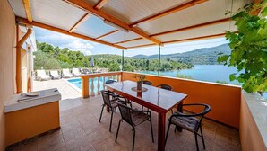 Un espacio al aire libre techado con una mesa y sillas, con vista a una piscina y un paisaje pintoresco de colinas y agua.