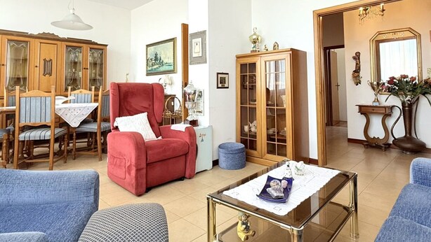 Una sala de estar con un sillón rojo, sofás azules, una mesa de centro de vidrio, armarios de madera y un comedor con mesa y sillas.