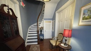 Eine Treppe, die zu einer zweiten Etage führt, ein Holzschrank, ein Stuhl, ein Tisch mit einer Lampe und ein gerahmtes Bild an der Wand.
