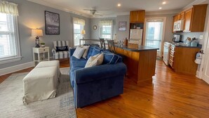 Ein geräumiges Wohnzimmer mit einem blauen Sofa, einem kleinen weißen Hocker und einer Küchenzeile mit Holzschränken und einer Mikrowelle.