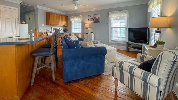 Ein geräumiges Wohnzimmer mit einem blauen Sofa, einem gestreiften Sessel und einem Fernseher. Im Hintergrund ist eine Küchenzeile mit Holzschränken und einer Bar erkennbar.