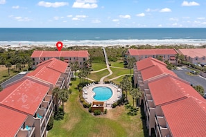 A resort with a central swimming pool, beachfront view, and multiple buildings with red roofs.