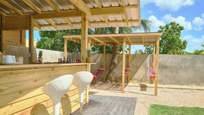 Eine hölzerne Sitzgruppe im Freien mit zwei weißen Stühlen, einer Holz-Pergola und einer Betonwand mit Grünpflanzen im Hintergrund.