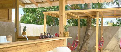 An outdoor wooden bar area with a metal roof, two white bar stools, a pineapple on the counter, and a hanging hammock.