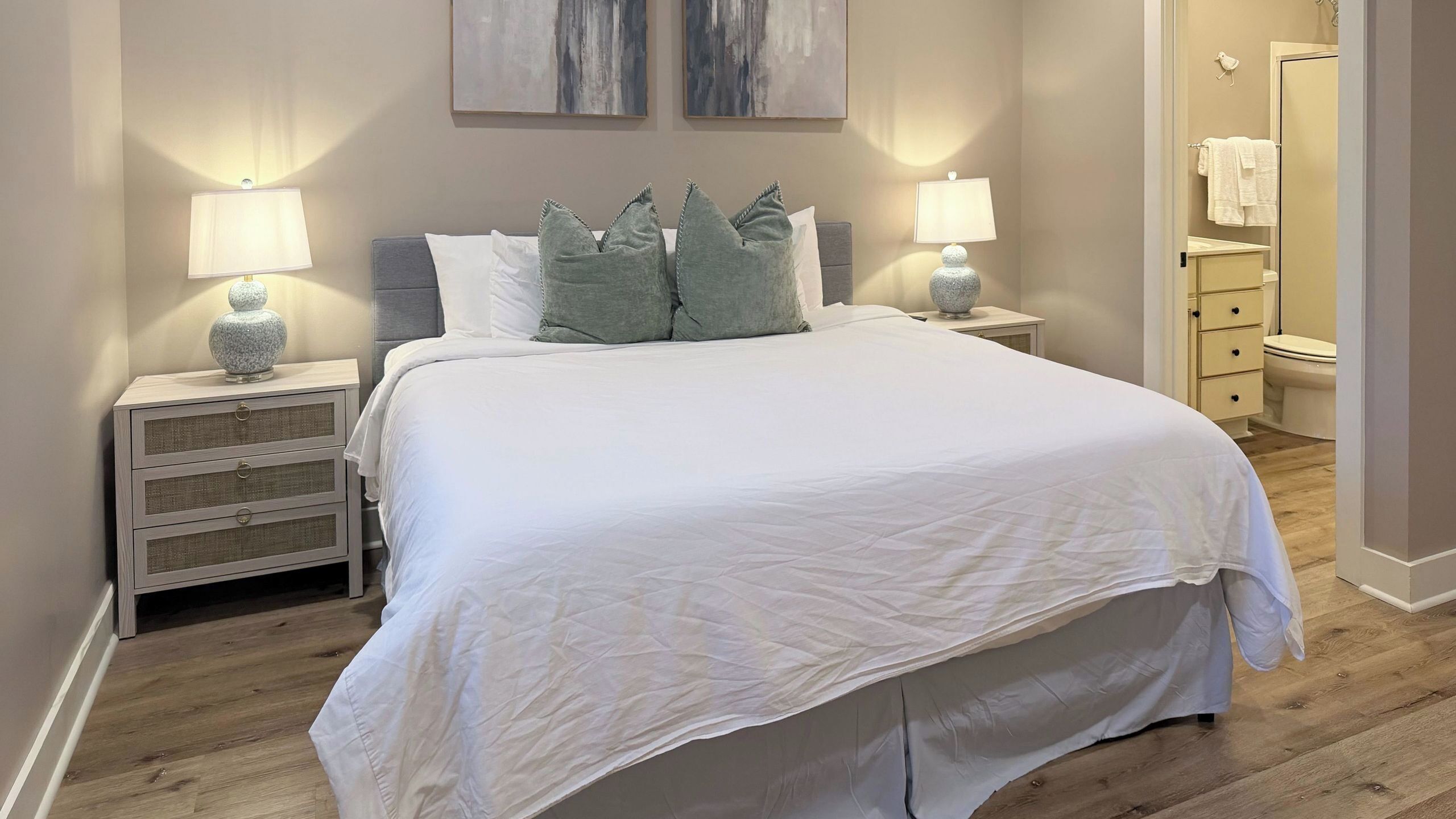 A neatly arranged bedroom with a large bed, two bedside tables with lamps, a nightstand, and a view of the bathroom.