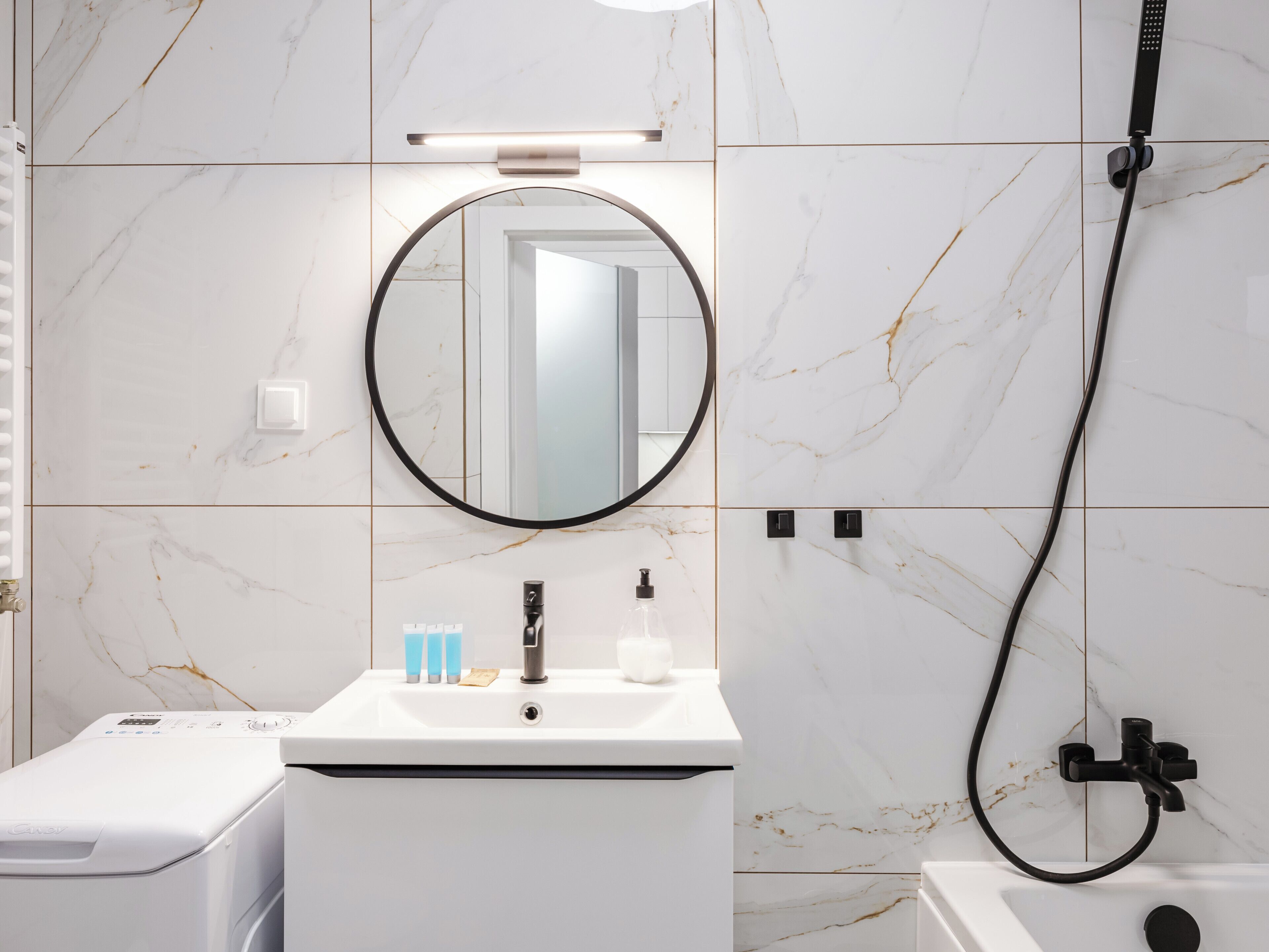 Un baño con lavamanos blanco, un espejo, ducha de mano y lavadora.