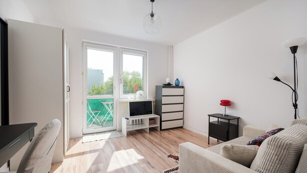 Ett modernt vardagsrum med en soffa, en tv och ett litet bord. Det finns en skjutdörr i glas som leder ut till en balkong med en stol.