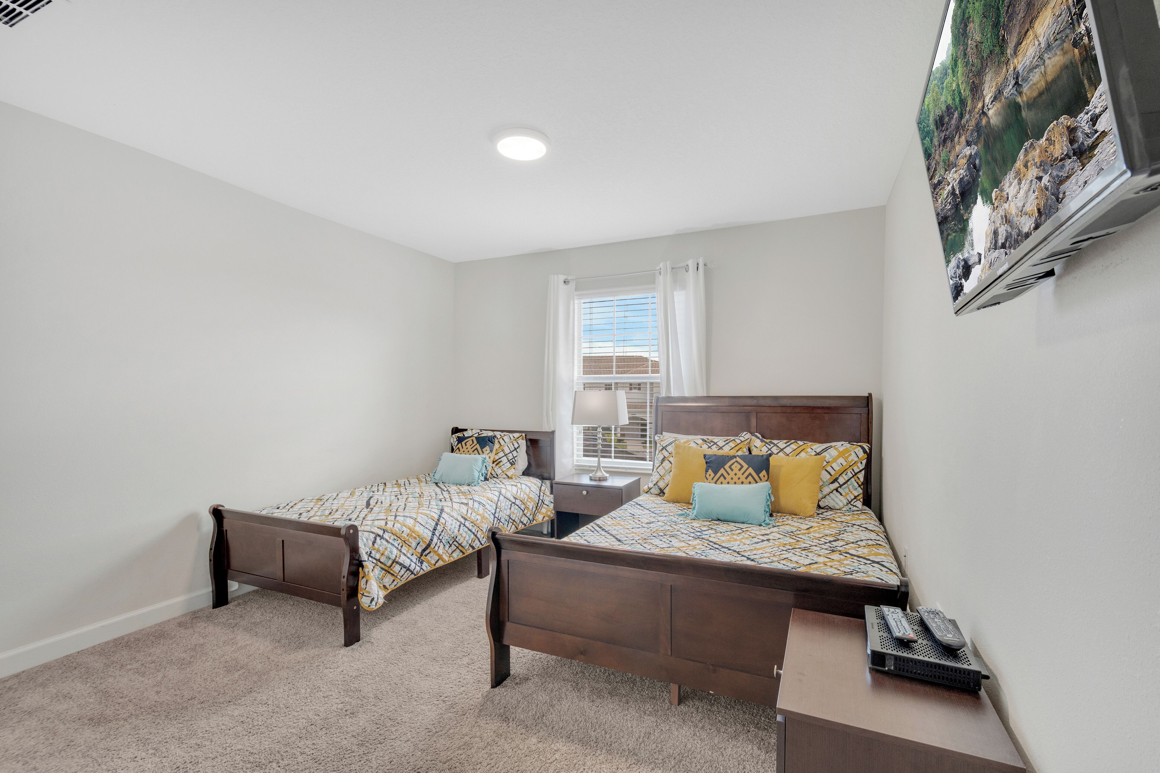 A bedroom with two single beds, a nightstand, a TV mounted on the wall, and a window with curtains.