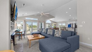 A spacious living area with a large blue sectional sofa, a wooden coffee table, and a dining area with a table and chairs. - Dune Fabulous | 1,200 ft to the beach | Private Pool, Hot Tub, and Dog Friendly (Corolla)
