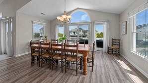 A spacious dining area with a large wooden table and chairs, a chandelier, and large windows offering a view of the neighborhood. - Dune Fabulous | 1,200 ft to the beach | Private Pool, Hot Tub, and Dog Friendly (Corolla)