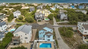 An aerial view of a coastal residential area with houses, a swimming pool, and a beach in the background. - Dune Fabulous | 1,200 ft to the beach | Private Pool, Hot Tub, and Dog Friendly (Corolla)