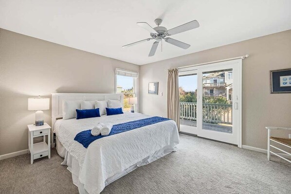 A bedroom with a large bed, a ceiling fan, a nightstand, a window, and a balcony.