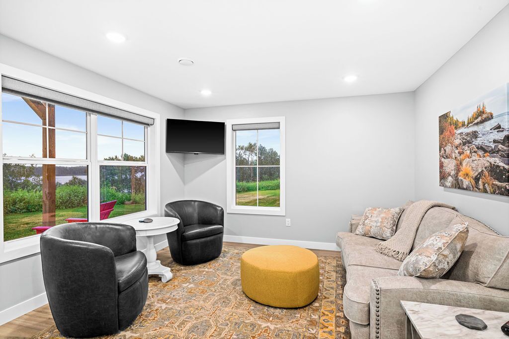 Un salon moderne comprenant un canapé, deux fauteuils, une table basse et un pouf jaune. De grandes fenêtres offrent une vue sur l’extérieur.