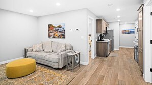 A modern apartment with a living room featuring a sofa, ottoman, and artwork, a kitchen with wooden cabinets, and a hallway leading to another room.