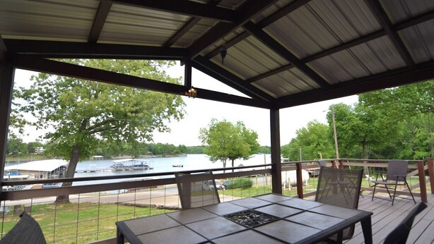 A covered outdoor patio with a table and chairs, overlooking a lake with boats.