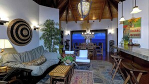 A cozy living room with a sofa, coffee table, and a chandelier. There is a dining area with a table and chairs, and a large window with a view.