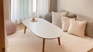 A modern living room with a white table, beige sofa, and a large window with sheer curtains.