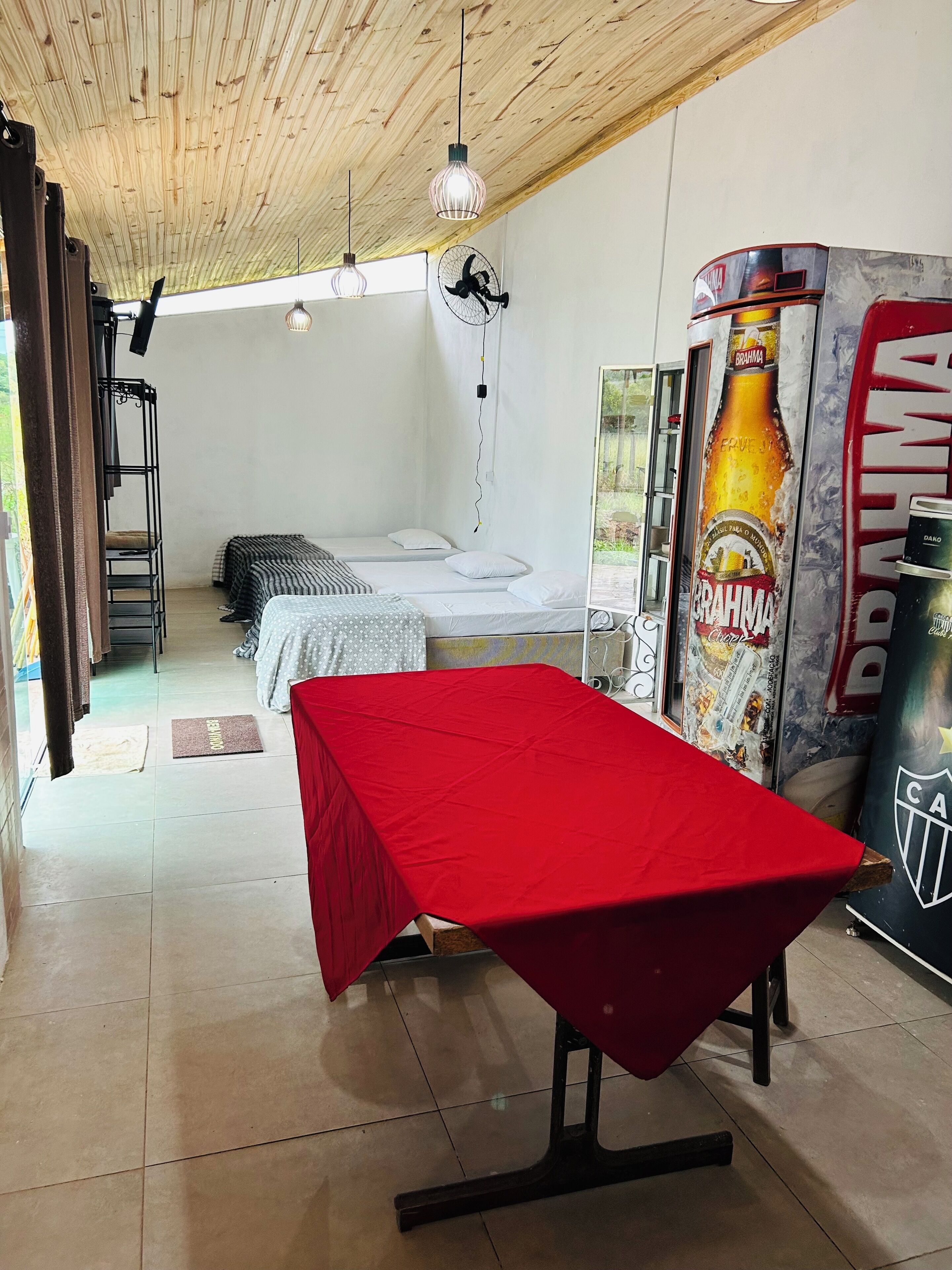 Una habitación con una mesa roja, literas, un ventilador y un anuncio grande de cerveza.