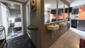 Une chambre d’hôtel avec un lit, une salle de bain dotée d’un grand miroir, un coin cuisine et un motif à carreaux sur un des murs.