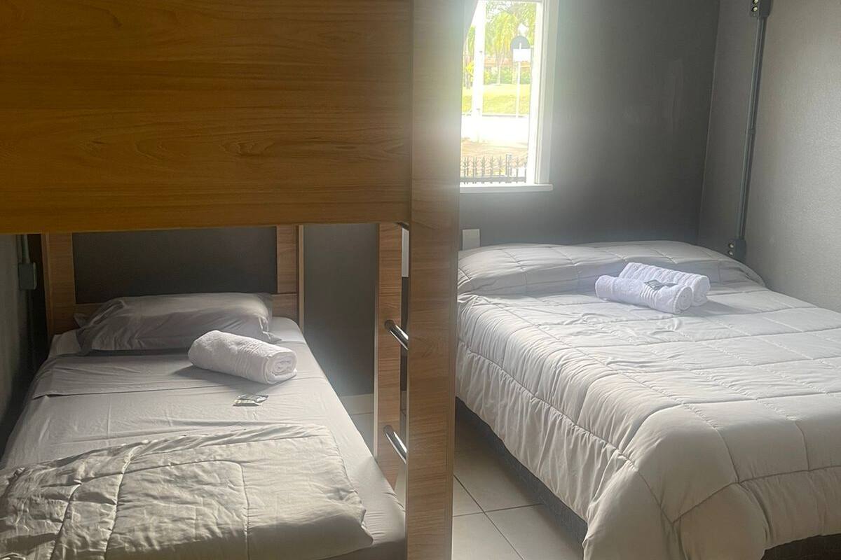 Ein Zimmer mit zwei Betten, eines mit einem Holz-Kopfteil und das andere mit einem Metallgestell. Ein Fenster mit Vorhängen, und der Boden ist mit Fliesen ausgelegt.