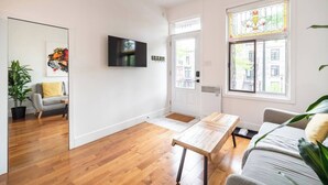 A modern living room with hardwood flooring, a flat-screen TV mounted on the wall, a large window with a stained glass design, and a door leading outside.