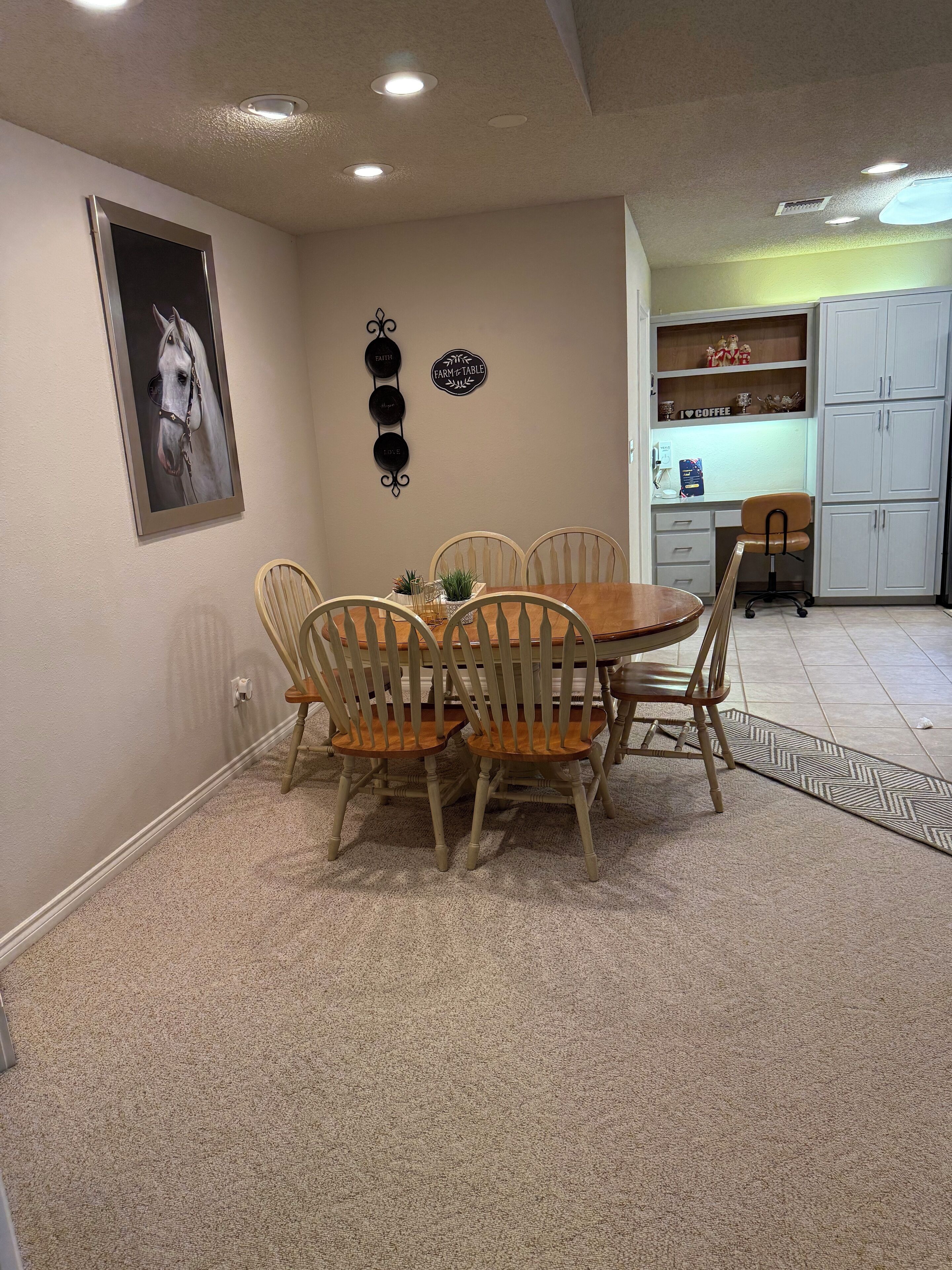 Un comedor con una mesa y sillas de madera, un cuadro de un caballo colgado en la pared, y una zona de cocina con armarios y encimera.