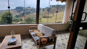 A room with a large window offering a view of a hilly landscape. There is a sofa and a small table with a glass container on it. - The Green House of Cunha (Sao Paulo)