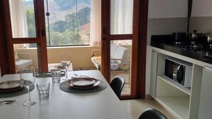 A dining area with a table set for two, a kitchen with a stove, and a view of a hillside through the window. - The Green House of Cunha (Sao Paulo)