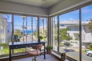Una habitación con escritorio, silla y ventanas amplias que dan a palmeras y edificios.