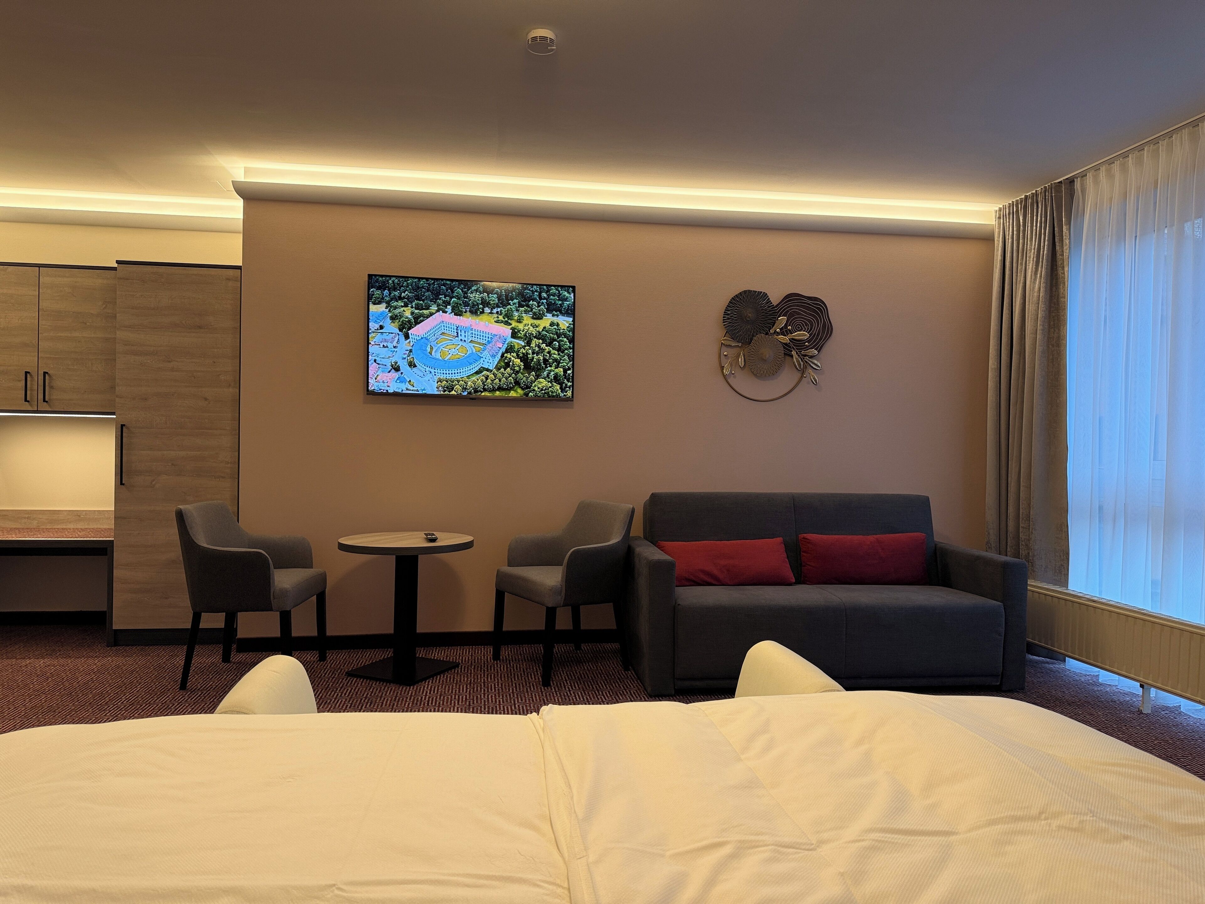 Une chambre d’hôtel comprenant un lit, un canapé gris avec des coussins rouges, deux fauteuils, une petite table ronde, un téléviseur à écran plat fixé au mur et un élément décoratif mural.