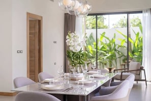 Un coin repas avec une longue table dressée pour six personnes, entourée de chaises, et une grande fenêtre donnant sur un espace extérieur verdoyant.