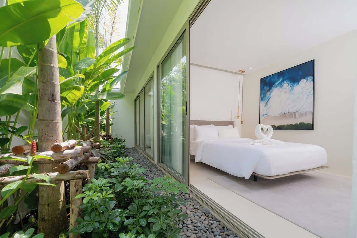 A modern hotel room with a large bed, a painting on the wall, and a view of a tropical garden.