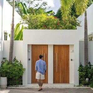 Um homem se aproxima de um edifício moderno com duas portas de madeira, cercado por vegetação exuberante e plantas tropicais.