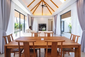 A spacious dining area with a wooden table and chairs, a ceiling fan, and large windows with curtains.
