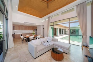 Un salon moderne avec un grand canapé d’angle, une table basse et un téléviseur. On y trouve également un coin repas avec une table et des chaises, et une cuisine à l’arrière-plan.