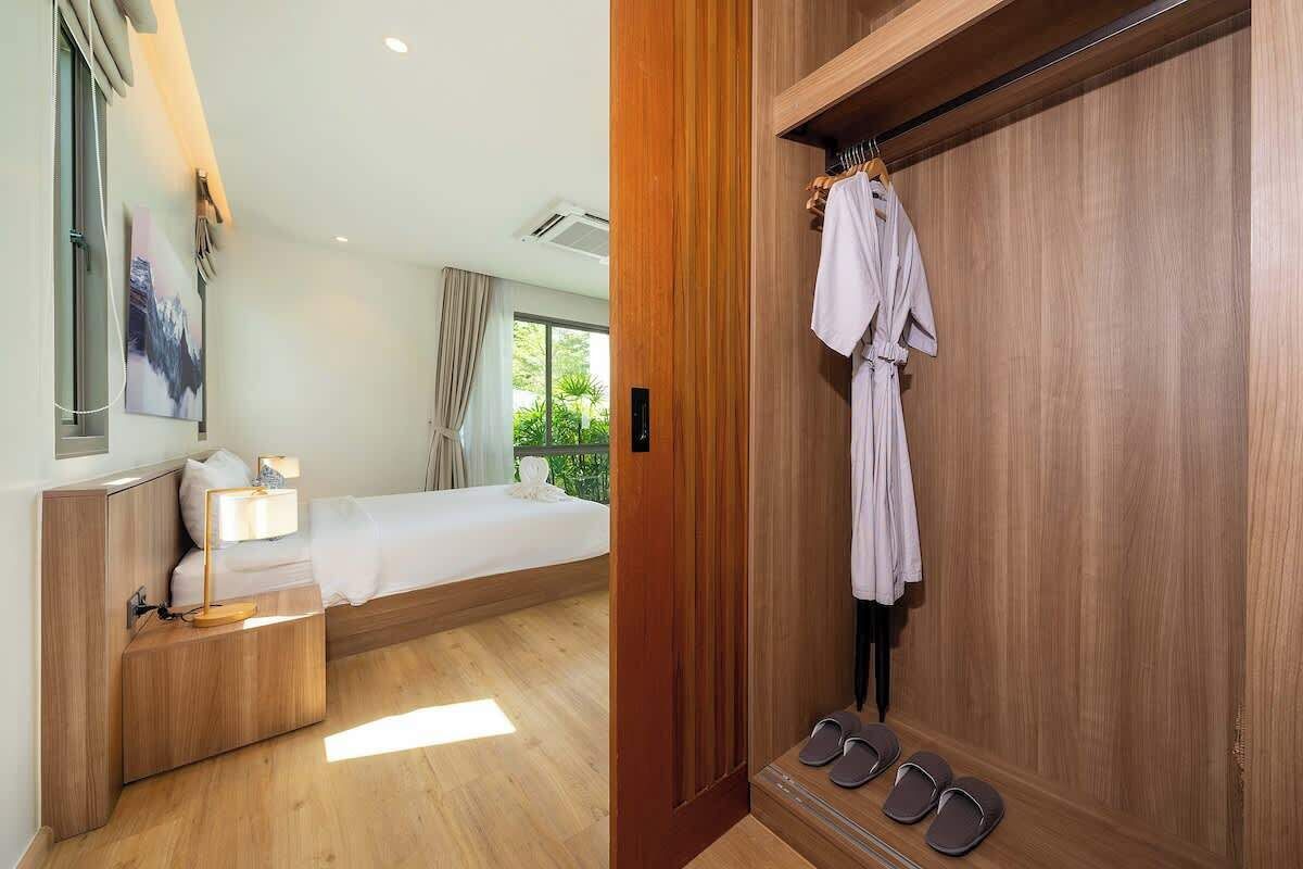 Une chambre d’hôtel comprenant un lit, une table de chevet, une lampe et une armoire en bois avec un peignoir et des chaussons.