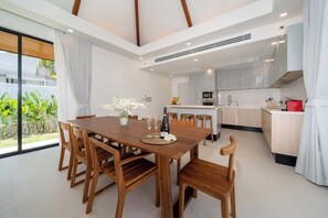 Une cuisine moderne avec des meubles blancs, un coin repas doté d’une table et de chaises en bois, et une grande fenêtre agrémentée de rideaux.